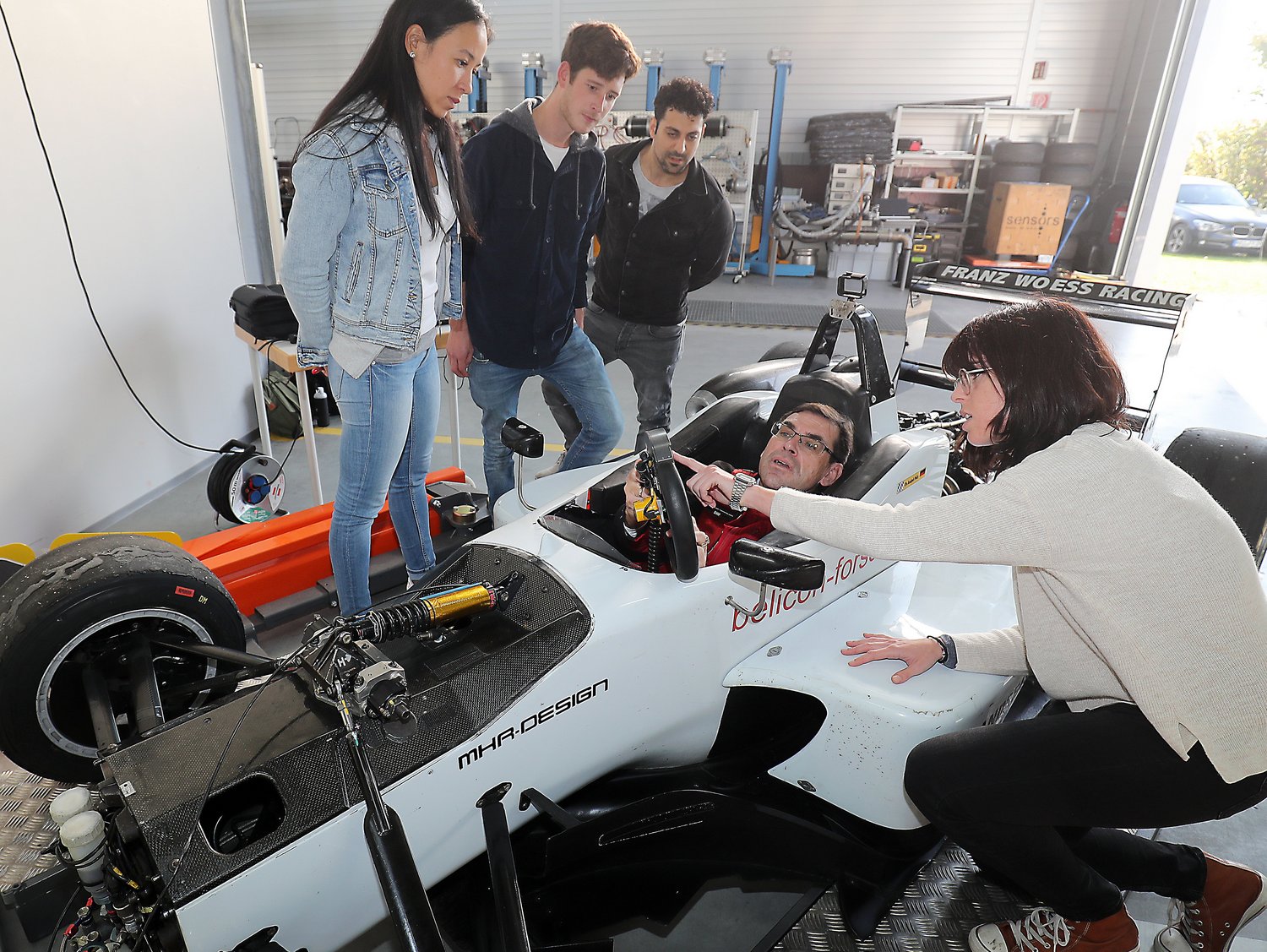 4 Studierende stehen an einem Formel 3-Rennwagen, der Dozent sitzt im Cockpit 