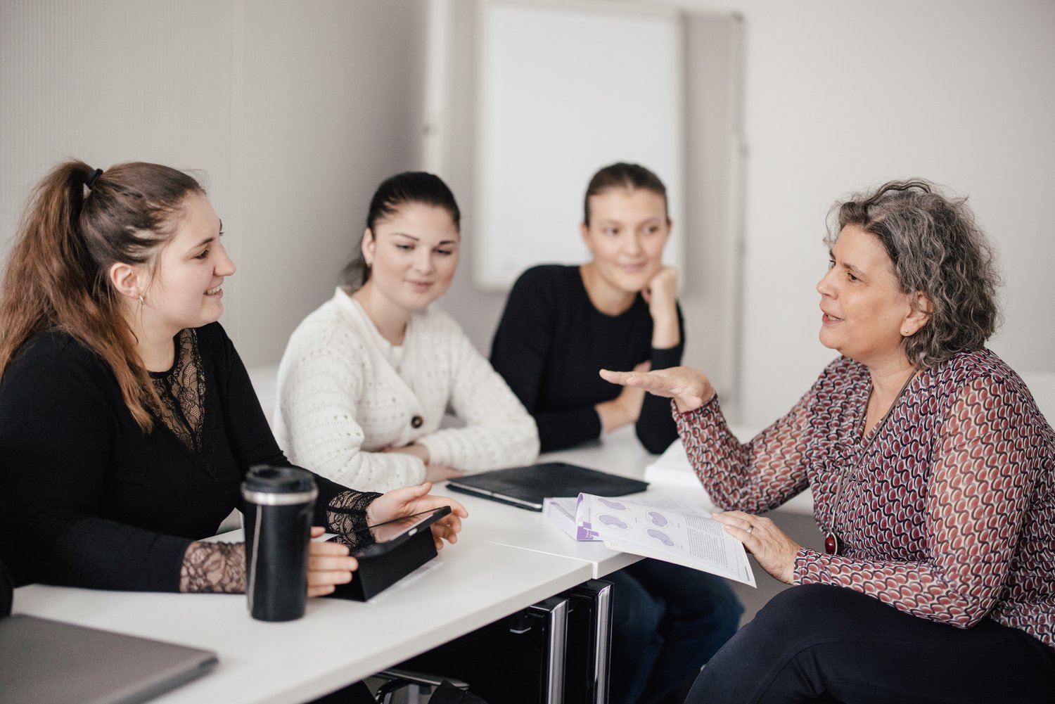 Eine Gruppe von vier Personen sitzt an einem Tisch. Eine Person erklärt etwas, während die anderen aufmerksam zuhören. Auf dem Tisch liegen Laptops und Unterlagen. Der Raum ist hell und modern gestaltet.
