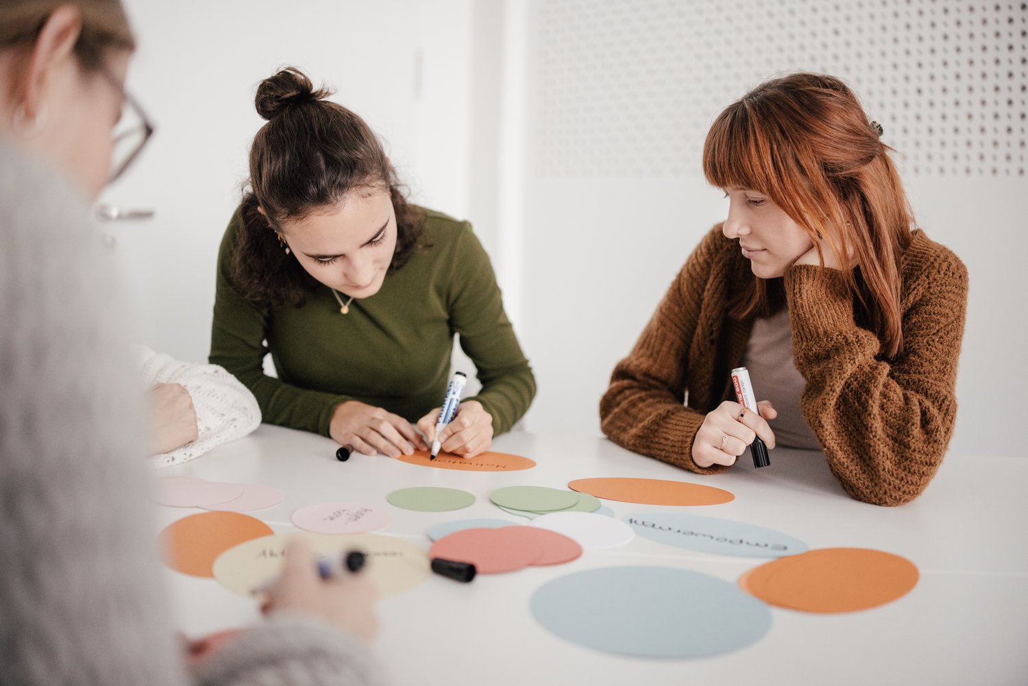 Eine Gruppe von Personen arbeitet an einem Tisch, umgeben von bunten Papierkarten. Eine Person schreibt mit einem Marker auf eine Karte, während eine andere aufmerksam zuschaut. Der Raum ist hell und modern gestaltet.