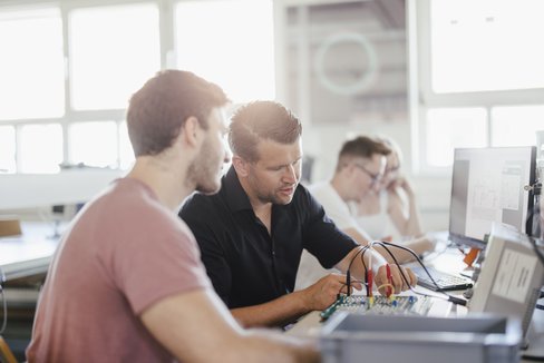 Zwei Personen sitzen nebeneinander, eine Person steckt Kabel an einen Schaltkreis an und die andere Person schaut zu.