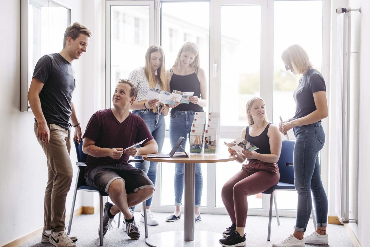 Man sieht sechs Personen in einem Raum. Zwei Personen sitzen an einem Tisch, die restlichen Personen stehen um den Tisch herum. Alle schauen sich Proschüren an.