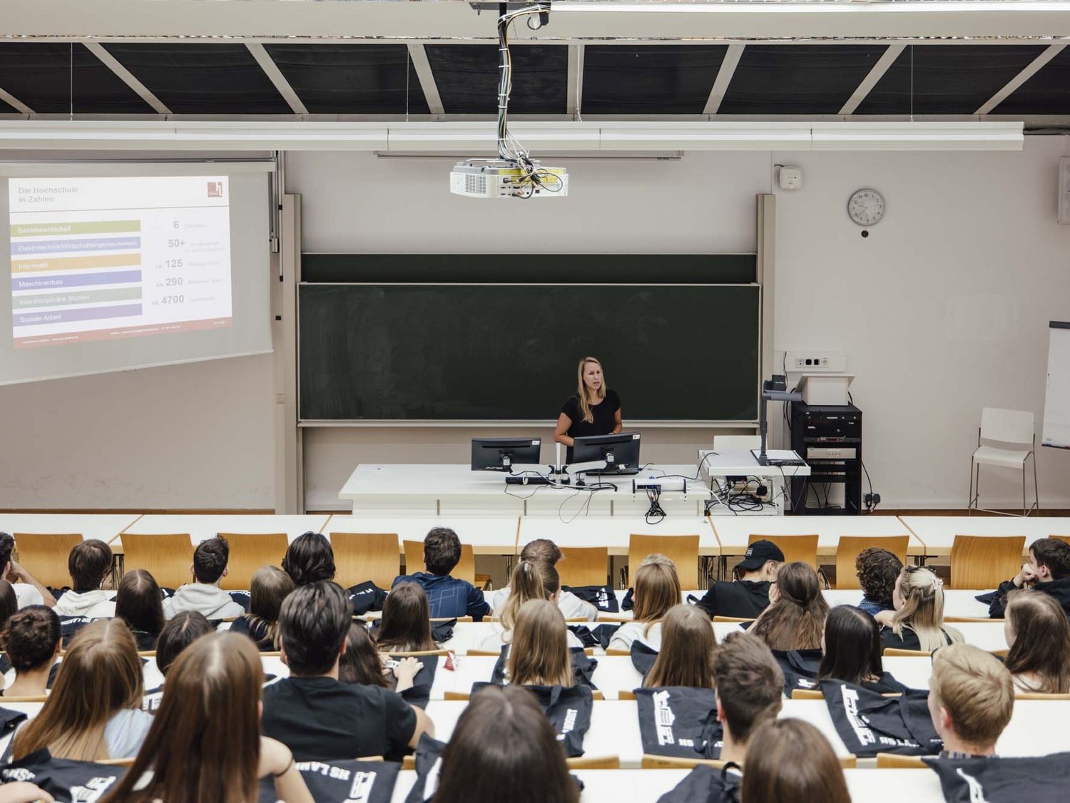 Man sieht mehrere sitzende Personen und eine Person die vorne im Raum an einer Tafel steht.