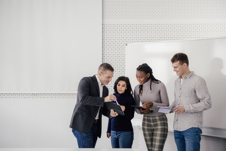 Eine Gruppe von vier Personen steht in einem modernen Raum. Eine Person zeigt auf ein Tablet, während die anderen aufmerksam schauen und Notizen machen. Alle wirken engagiert und in einem Austausch über ein Thema.