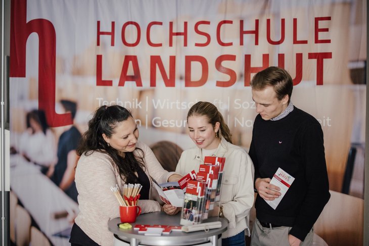 Drei Personen stehen an einem Tisch, der mit Informationsmaterialien zur Hochschule Landshut dekoriert ist. Im Hintergrund ist ein Banner mit dem Hochschulnamen und verschiedenen Fachrichtungen sichtbar.
