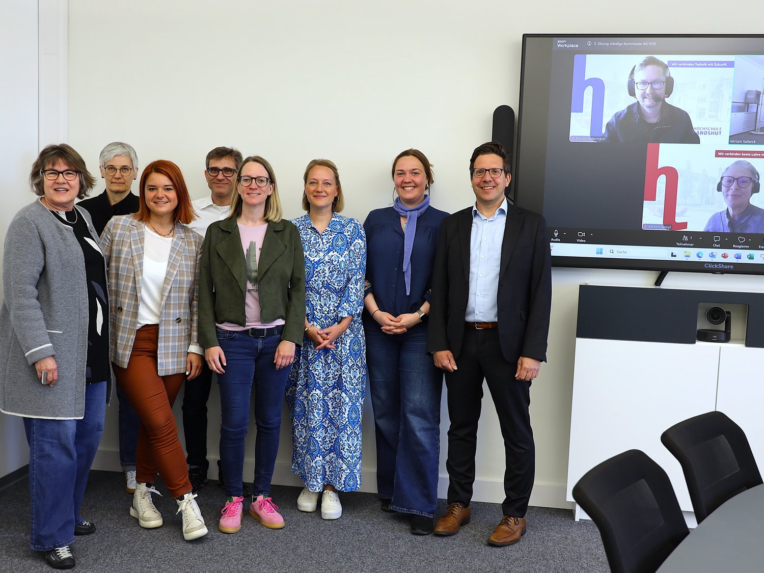 Zehn Teilnehmer der ständigen Kommission Kompass sind zu sehen, sie stehen nebeneinander und lächeln in die Kamera. Zwei der Mitglieder sind auf einem Bildschirm im Hintergrund zu sehen.