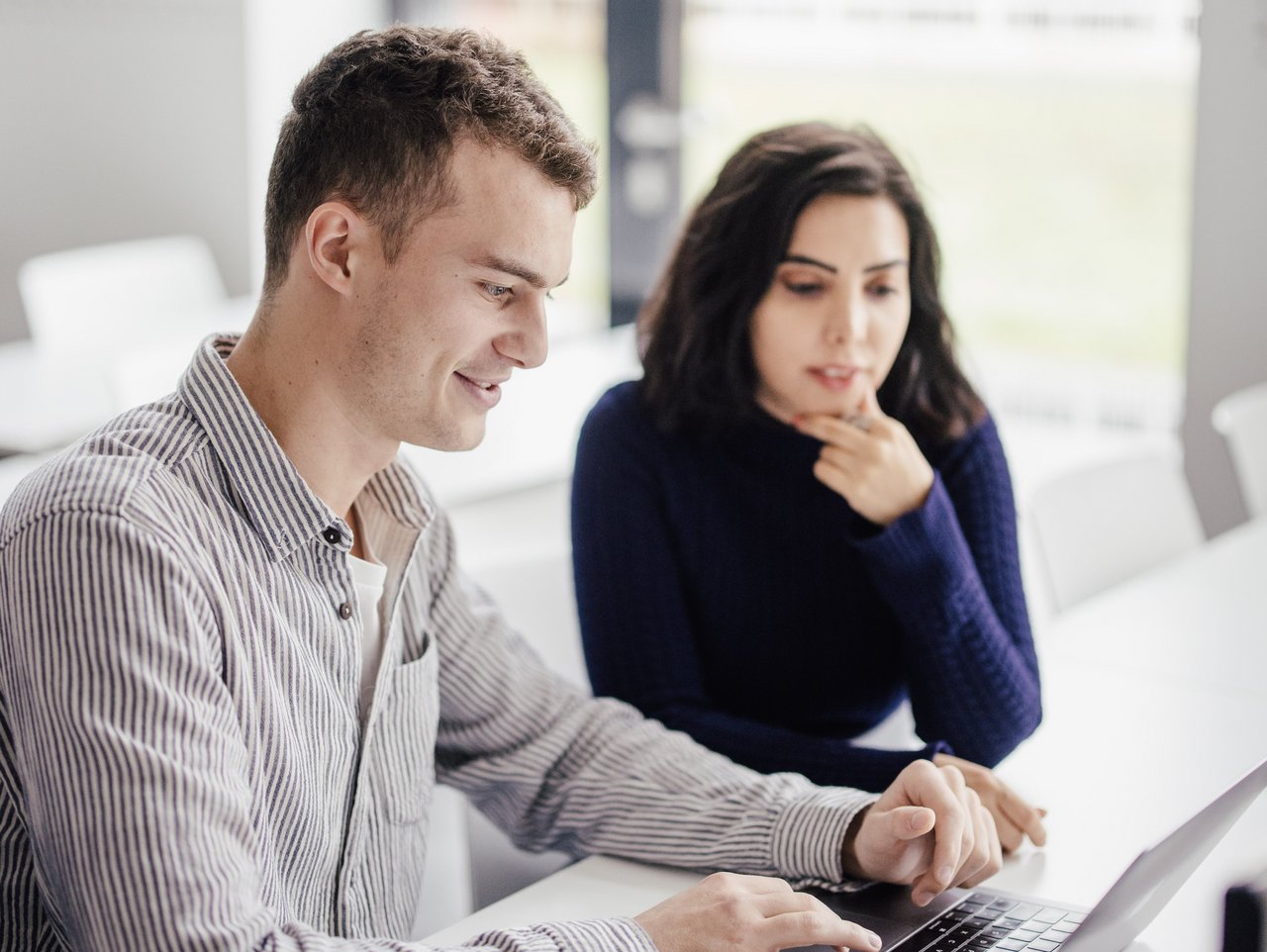 Zwei Personen sitzen an einem Tisch in einem modernen Raum. Eine Person arbeitet am Laptop, während die andere nachdenklich zuschaut. Helle Farben und große Fenster sorgen für eine freundliche Atmosphäre.