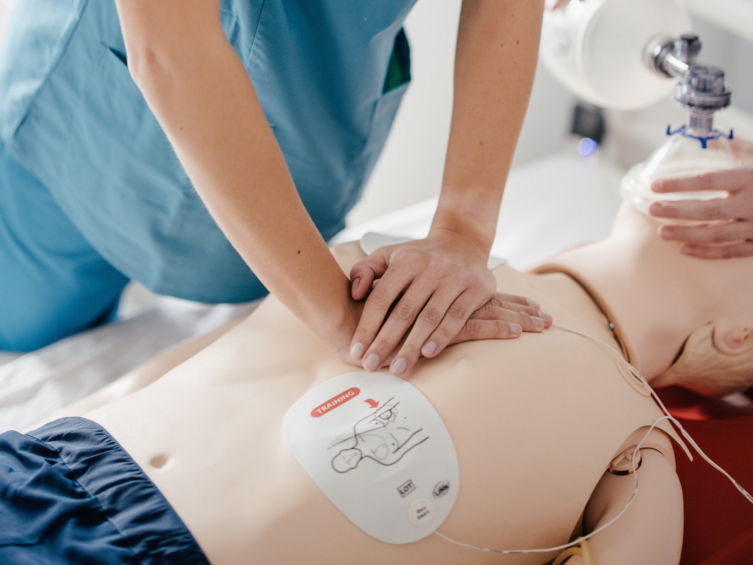 Eine Person in blauer medizinischer Kleidung führt eine Herz-Lungen-Wiederbelebung (HLW) an einer Übungspuppe durch. Im Hintergrund ist eine weitere Person mit einem Beatmungsgerät zu sehen.