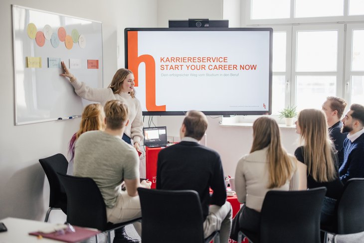 Eine Person präsentiert vor einer Gruppe von Studierenden in einem modernen Seminarraum. Auf einem Bildschirm wird der Karriereservice und der Slogan "Start your career now" angezeigt. Notizen und Materialien sind sichtbar.
