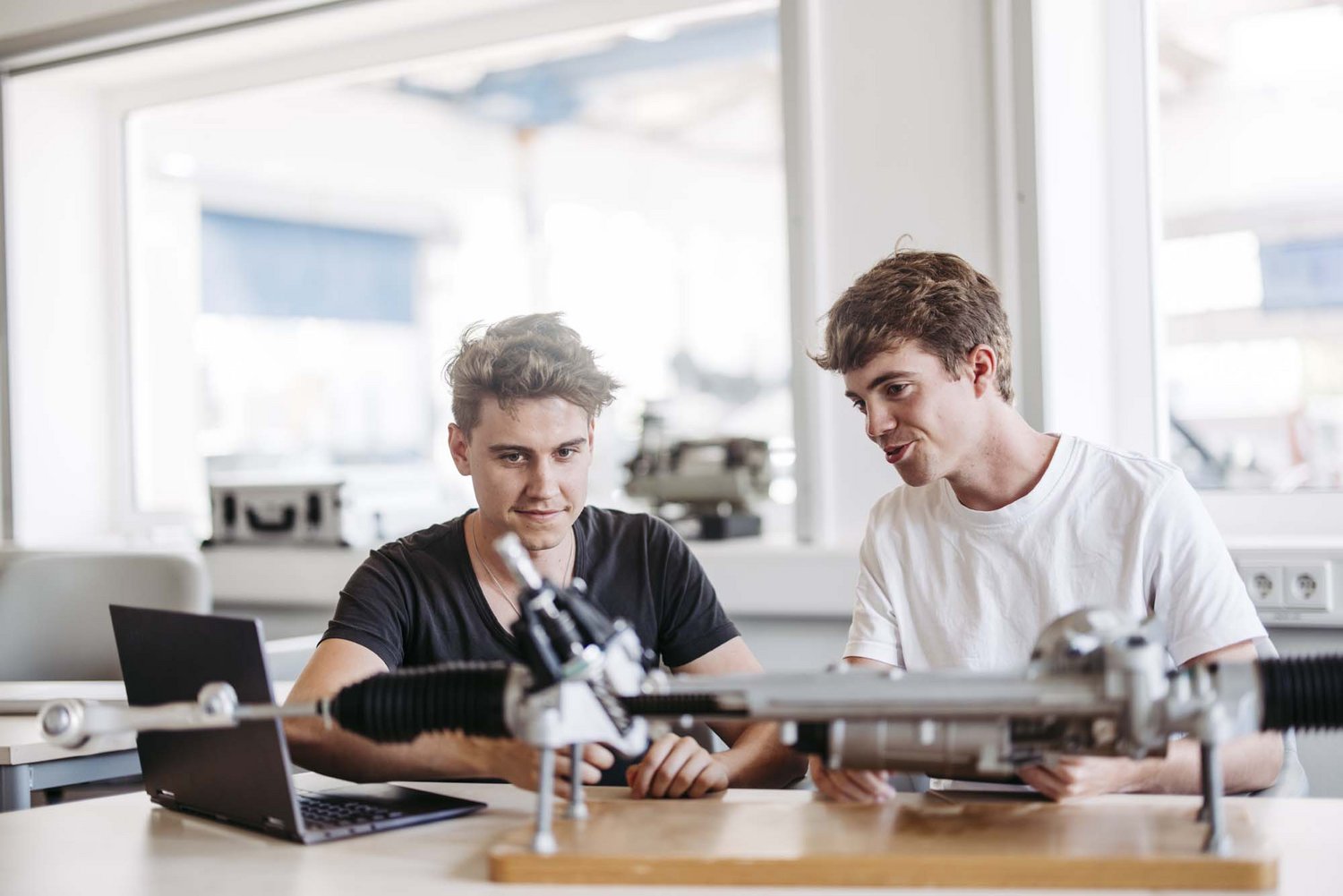 Zwei Studierende sitzen mit Laptop und Tablet und betrachten einen Getiebewellenaufaufbau