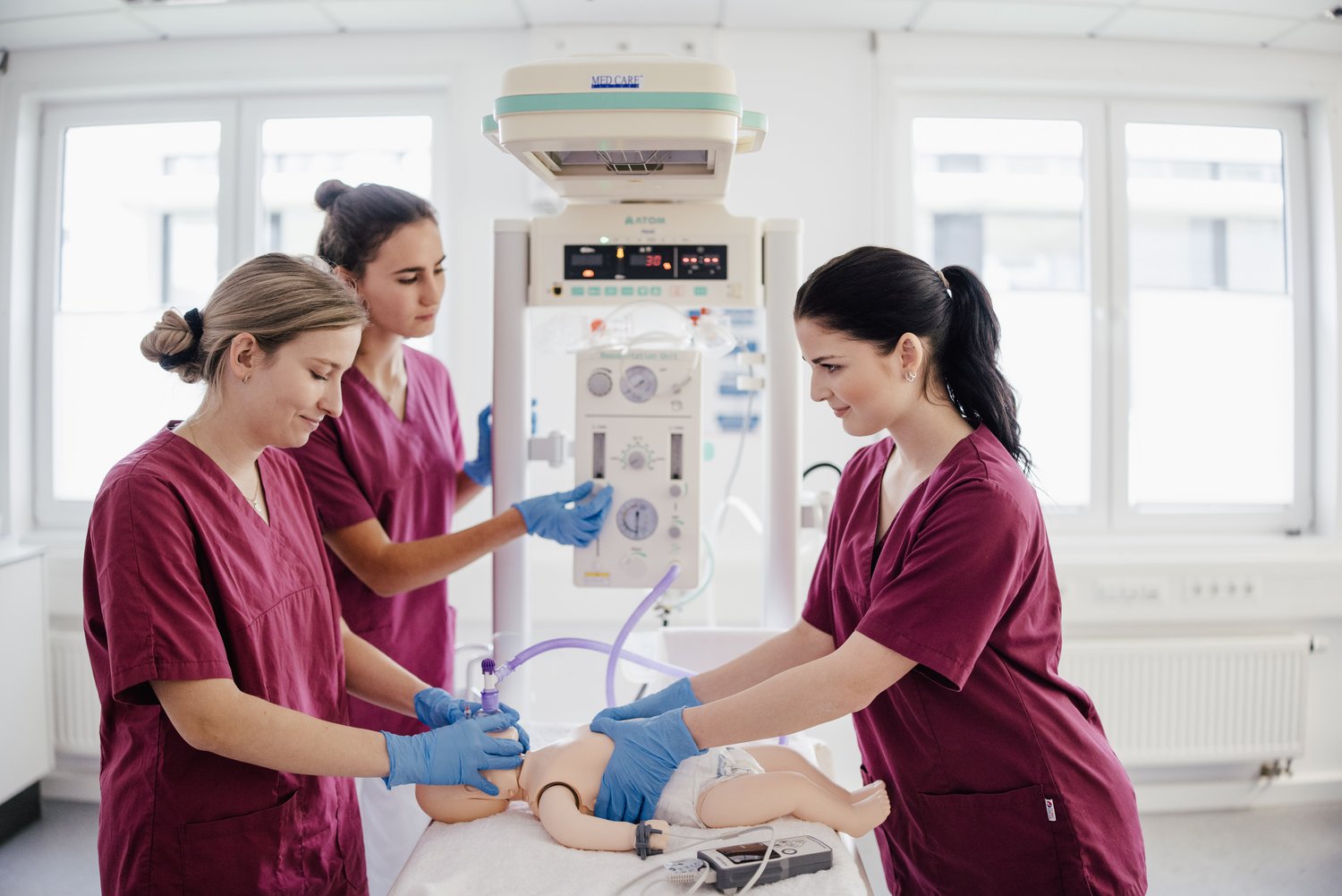 Drei Personen in medizinischer Kleidung arbeiten an einem Dummy in einem Krankenhaus. Sie verwenden Geräte zur Überwachung und Unterstützung der Atmung, während sie sich auf die Simulation einer medizinischen Notfallsituation konzentrieren.