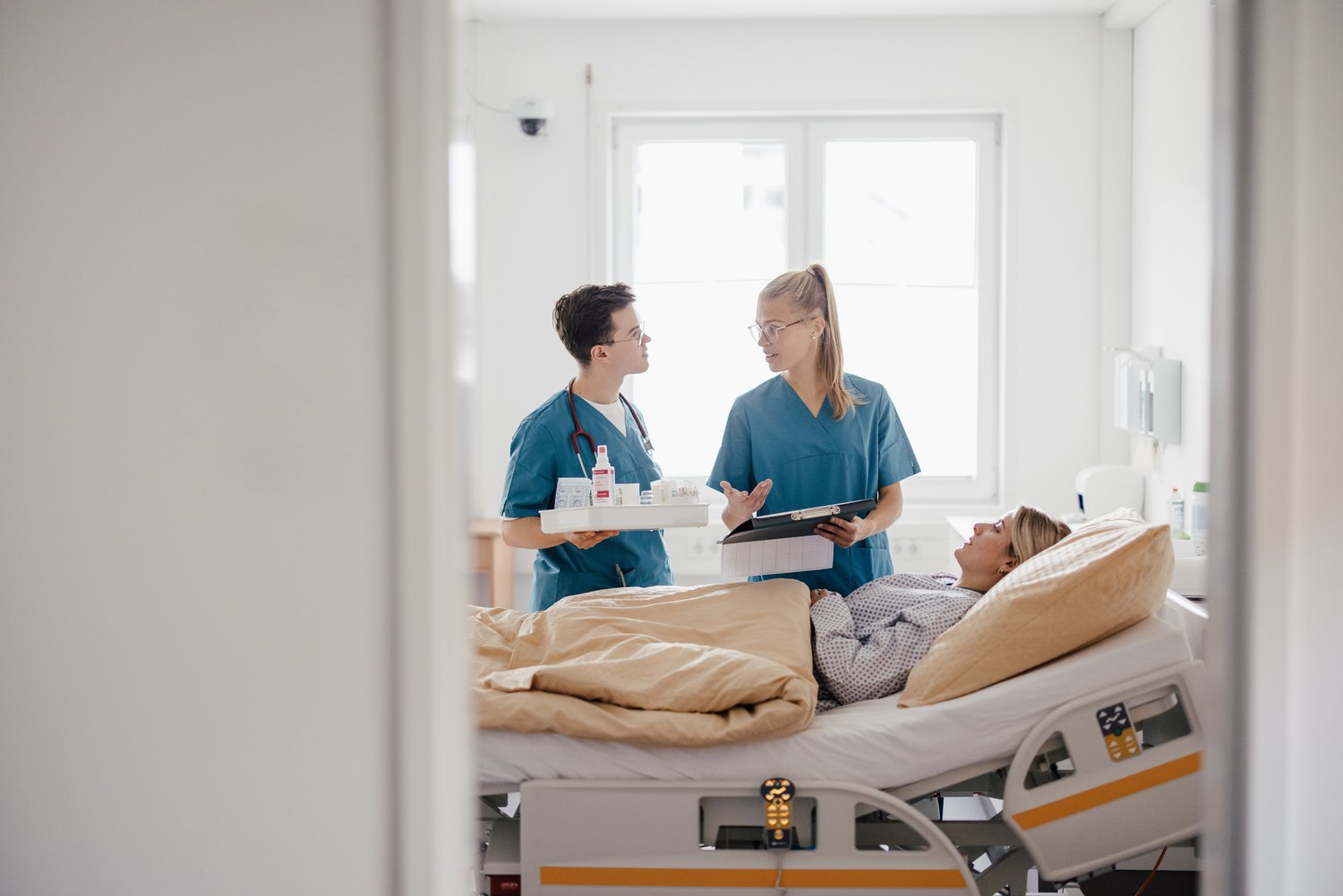 Zwei Pflegekräfte in blauen Scrubs stehen in einem Krankenhauszimmer und sprechen miteinander, während eine Patientin im Bett liegt. Auf einem Nachttisch sind medizinische Utensilien sichtbar. Tageslicht strömt durch das Fenster.