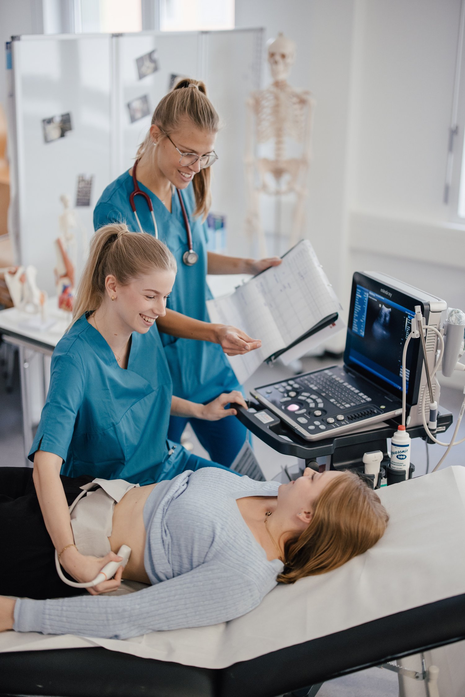 Eine Person liegt auf einer Liege und erhält eine Ultraschalluntersuchung. Zwei weitere Personen, in medizinischer Kleidung, stehen daneben und beobachten den Bildschirm sowie die Ergebnisse. Im Hintergrund sind anatomische Modelle sichtbar.