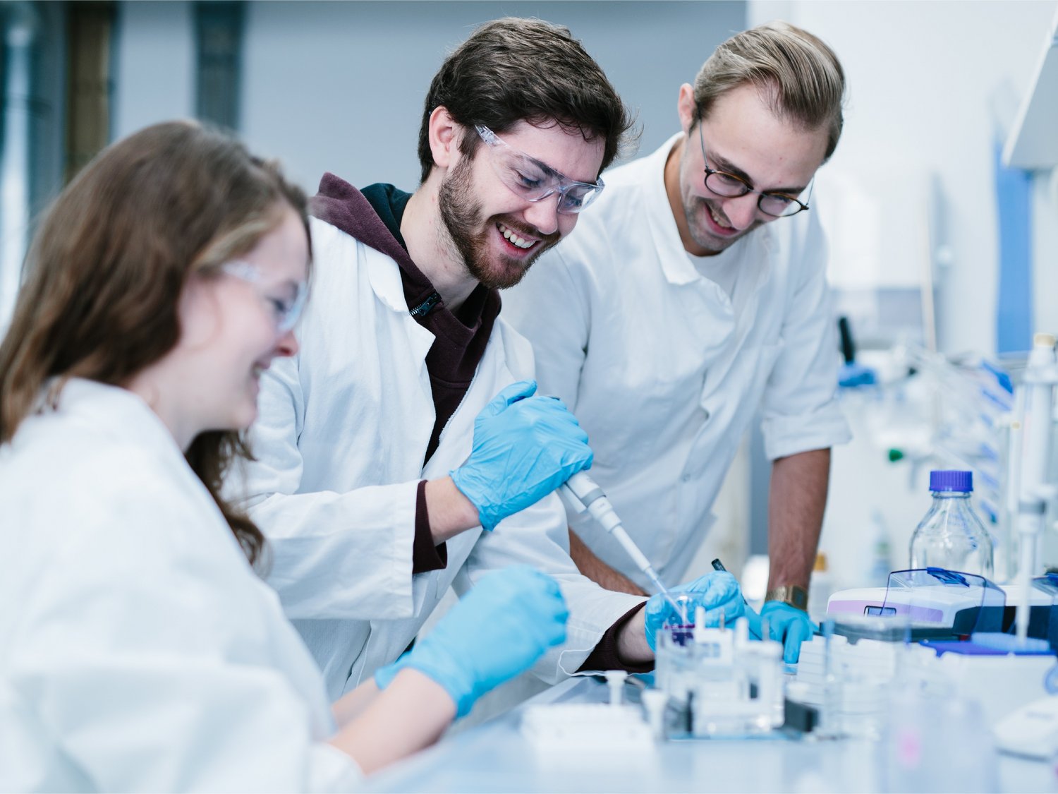 Drei Studierende stehen an einem chemischen Laborarbeitsplatz. Einer von ihnen benutzt eine Pipette.