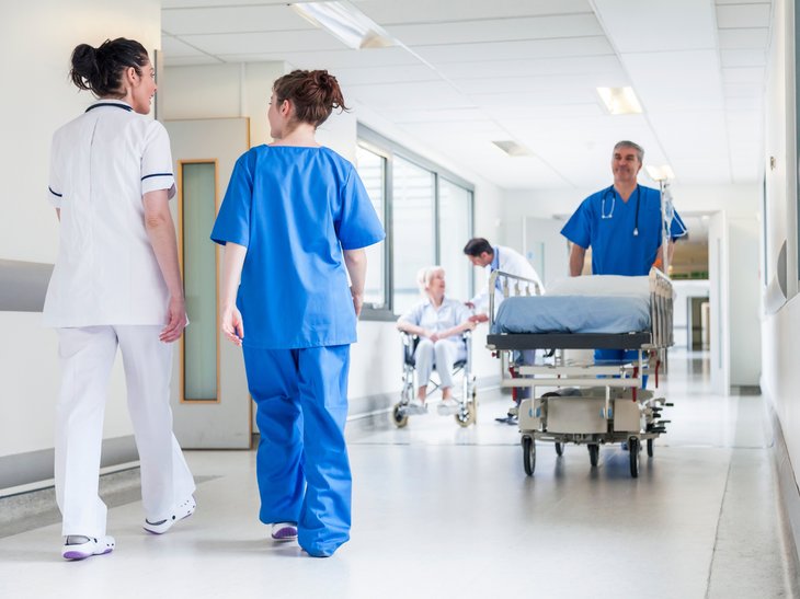 Mehrere Personen in einem Klinikflur sind zu sehen. Zwei Personen reden jeweils miteinander, eine schiebt ein Bett.