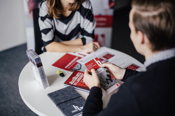 Eine Person sitzt an einem Tisch und hält ein Informationsblatt in der Hand. Eine andere Person ist im Hintergrund sichtbar. Auf dem Tisch liegen weitere Materialien und ein Notizbuch. Der Raum wirkt modern und einladend.