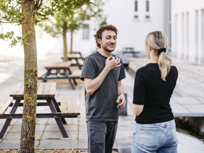 Zwei Personen stehen draußen nebeneinander die eine Person übt Gebärdensprache aus die andere Person schaut die eine Person an