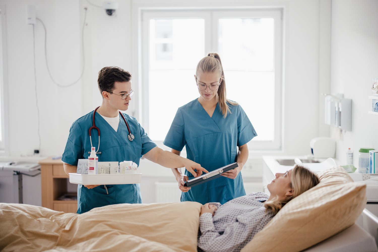 Zwei medizinische Fachkräfte in blauen Scrubs stehen neben einem Bett. Eine Person im Bett schaut aufmerksam auf ein Tablet, während die andere auf den Bildschirm zeigt. Im Hintergrund sind Fenster und medizinische Geräte sichtbar.