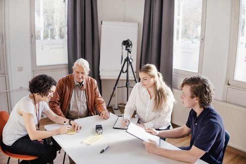 Eine Gruppe von vier Personen sitzt an einem Tisch in einem hellen Raum. Sie diskutieren und notieren Informationen. Im Hintergrund steht ein Stativ mit einer Kamera. Notizblätter und ein Aufnahmegerät liegen auf dem Tisch.