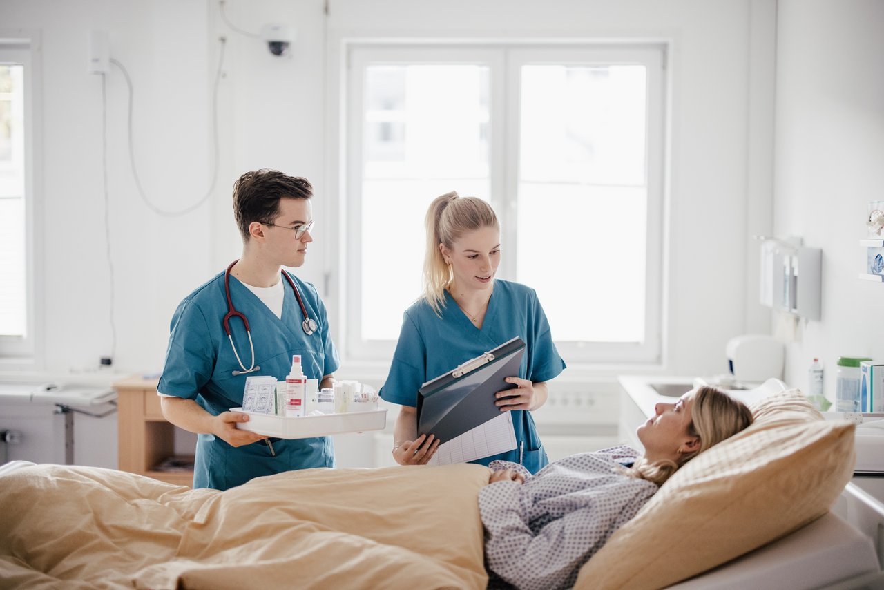 Ein medizinisches Team in blauen Scrubs steht neben einem Bett. Eine Person hält eine Tablett mit Medikamenten, während die andere eine Patientenakte zeigt. Eine Patientin liegt im Bett und hört aufmerksam zu.