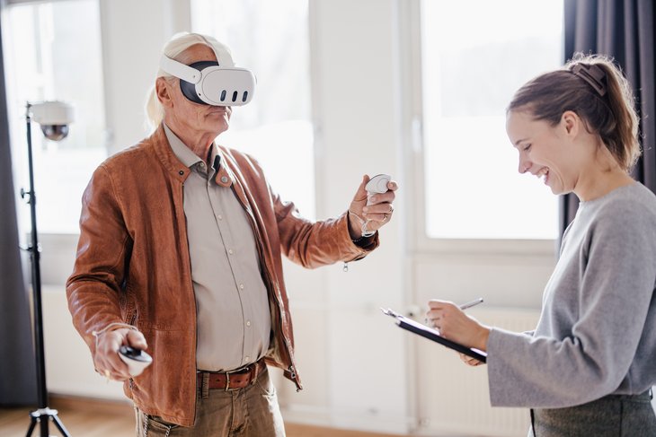 Eine Person trägt eine VR-Brille und hält einen Controller in der Hand, während eine andere Person lächelnd Notizen macht. Der Raum ist hell und modern eingerichtet, mit Fenstern im Hintergrund.
