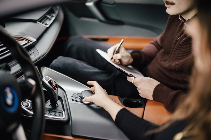 Eine Person sitzt im Fahrersitz eines BMW und hält einen Stift sowie ein Notizbuch. Eine andere Person greift nach dem Bedienelement in der Mittelkonsole. Der Innenraum des Fahrzeugs ist modern und stilvoll gestaltet.