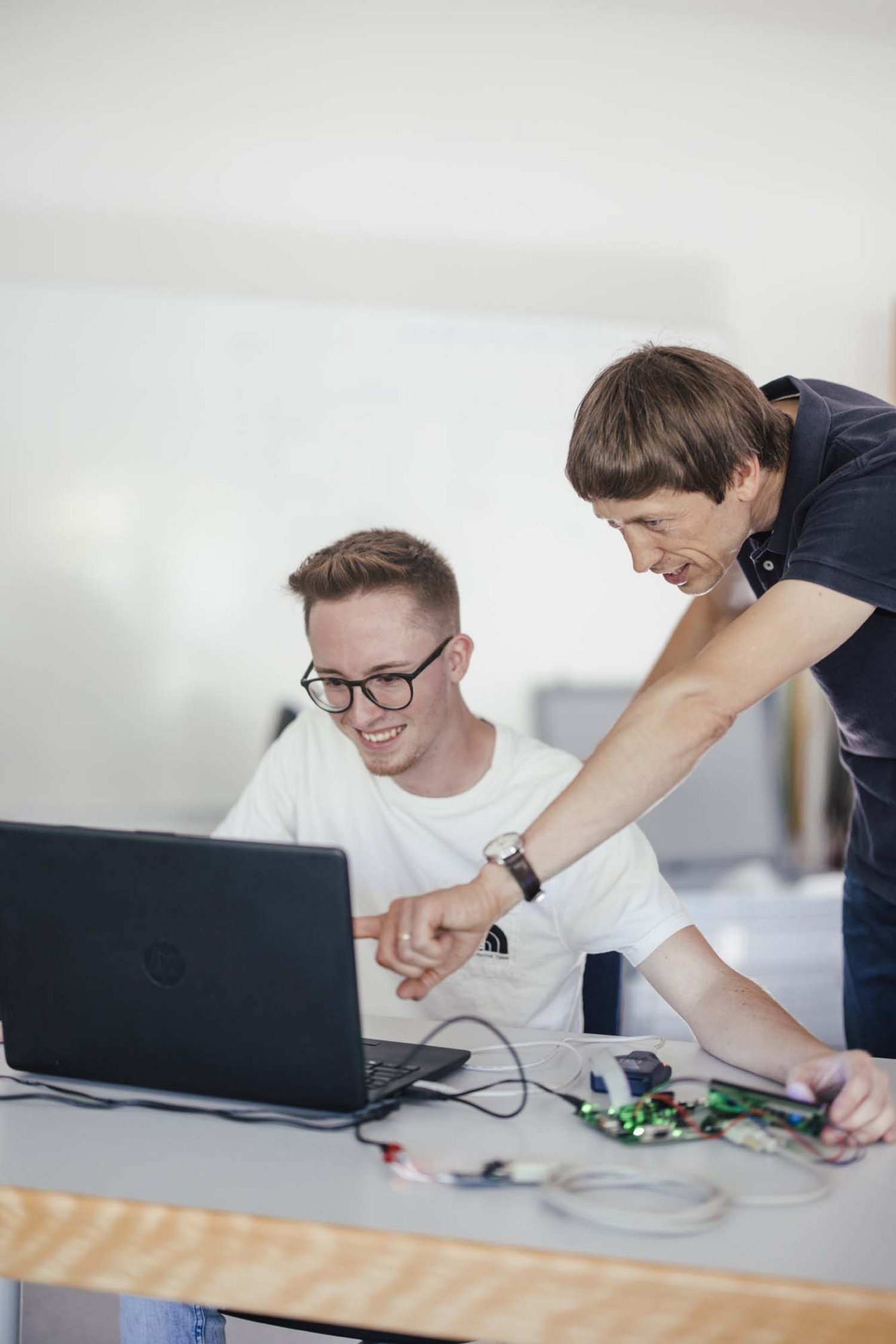 Zwei Personen sind zusehen. Vor ihnen steht ein Computer dieser ist angeschlossen an irgendwelche Schaltkreise. Die eine Person sitzt die andere steht. Die stehende Person zeigt mit dem Arm Richtung Bildschirm