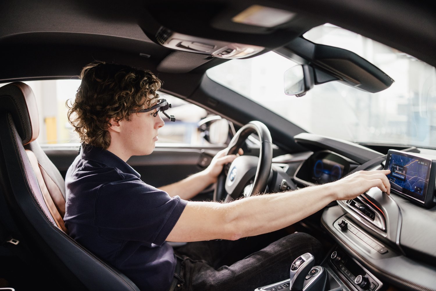 Eine Person sitzt im Fahrersitz eines Autos und bedient das Infotainmentsystem. Die Person trägt eine spezielle Brille und konzentriert sich auf den Bildschirm, während sie das Lenkrad hält.