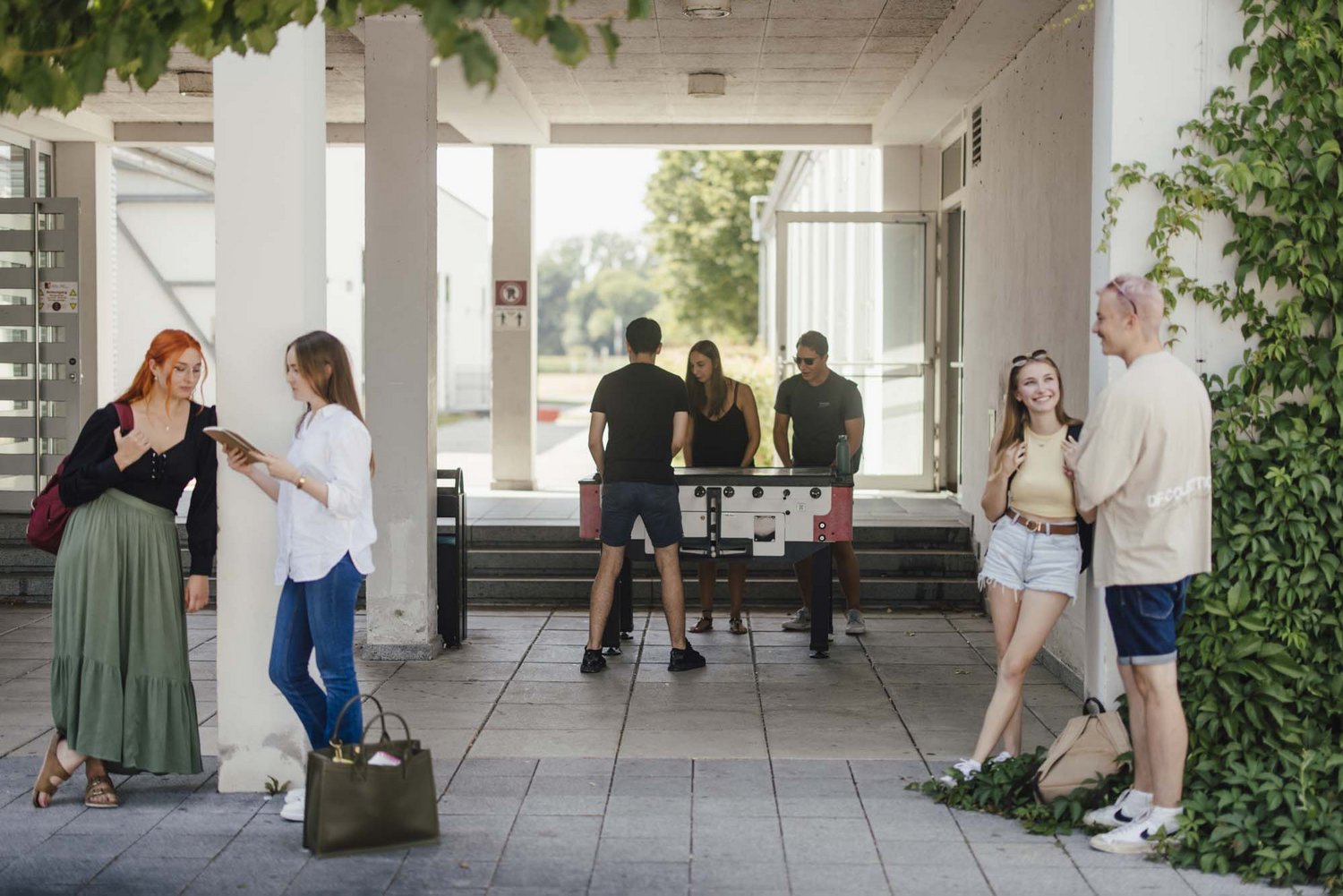 Studierende unterhalten sich draußen. Im Hintergrund spielt eine Gruppe am Kicker