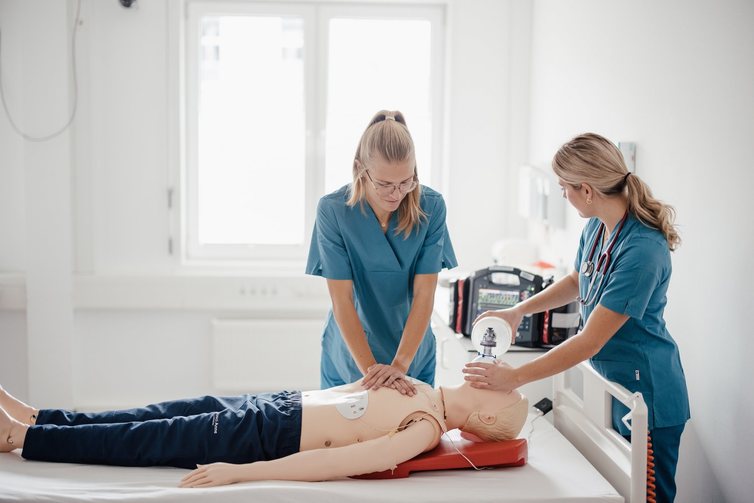 Zwei Personen in blauen medizinischen Kitteln führen eine CPR-Übung an einer Übungspuppe durch. Eine Person führt Herzkompressionen durch, während die andere eine Beatmungsmaske anlegt. Der Raum ist hell und modern.