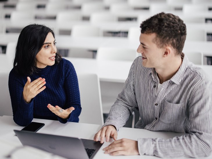 Zwei Personen sitzen an einem Tisch in einem modernen Raum. Eine Person spricht gestikulierend, während die andere aufmerksam zuhört. Auf dem Tisch steht ein Laptop und ein Smartphone. Im Hintergrund sind leere Stühle zu sehen.