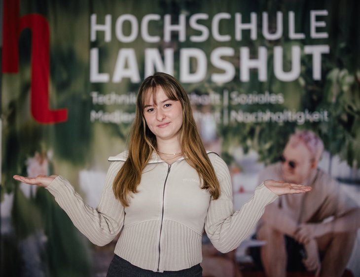 Eine Person steht vor einem Banner der Hochschule Landshut, das Themen wie Technik, Soziales, Medien und Nachhaltigkeit hervorhebt. Die Person hat lange Haare und trägt einen hellen Pullover, während sie die Hände offen präsentiert.