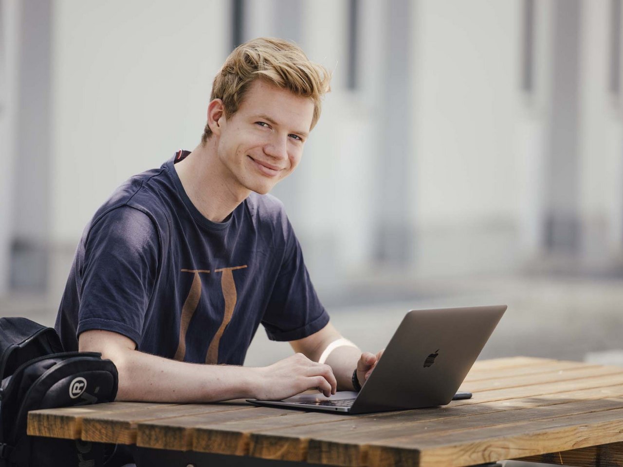 Eine Person sitzt draußen, lächelt und hat ihren Pc aufgeklappt.