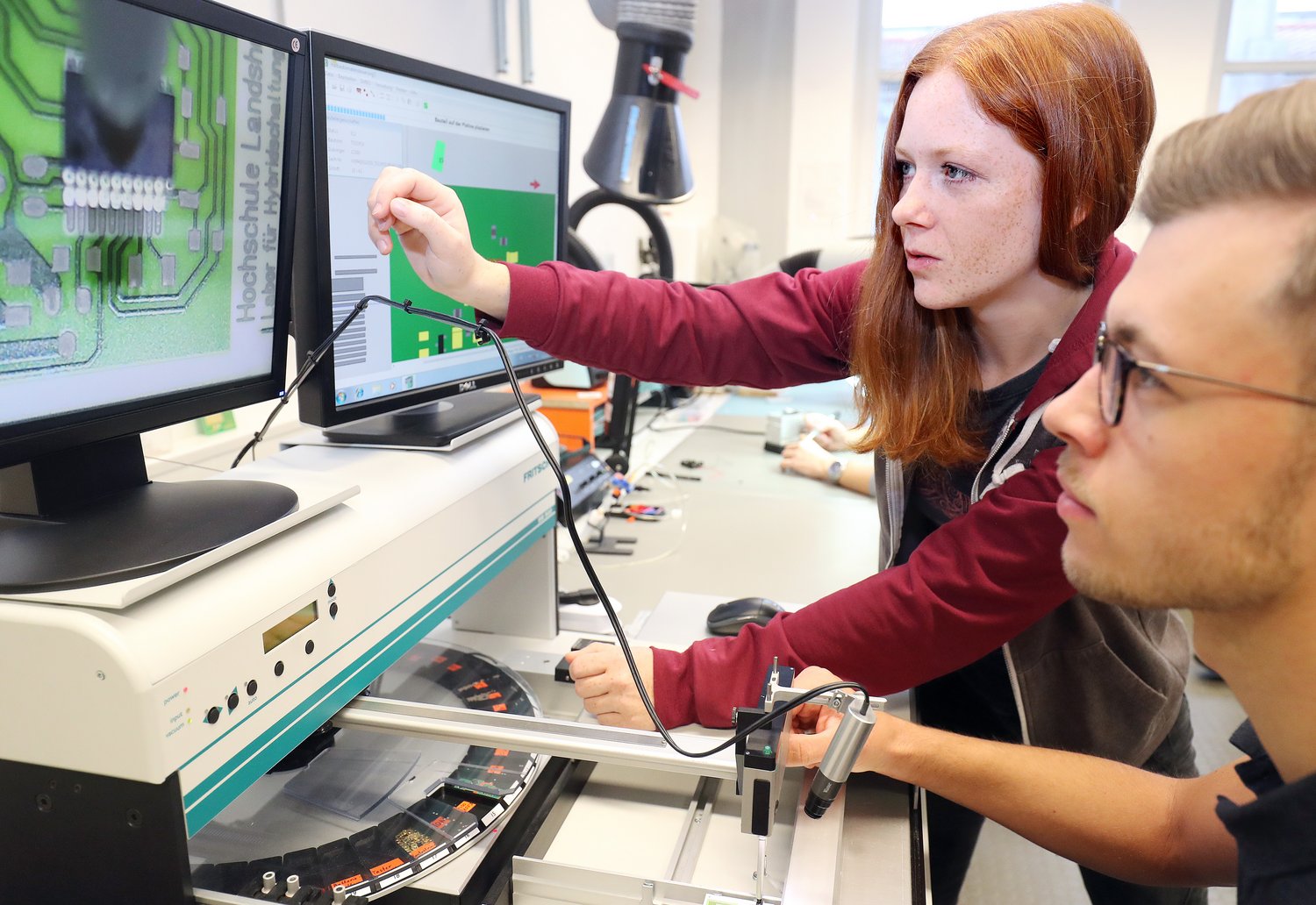 Zwei Personen sitzen im Physiklabor vor einem Gerät mit Bildschirmen.