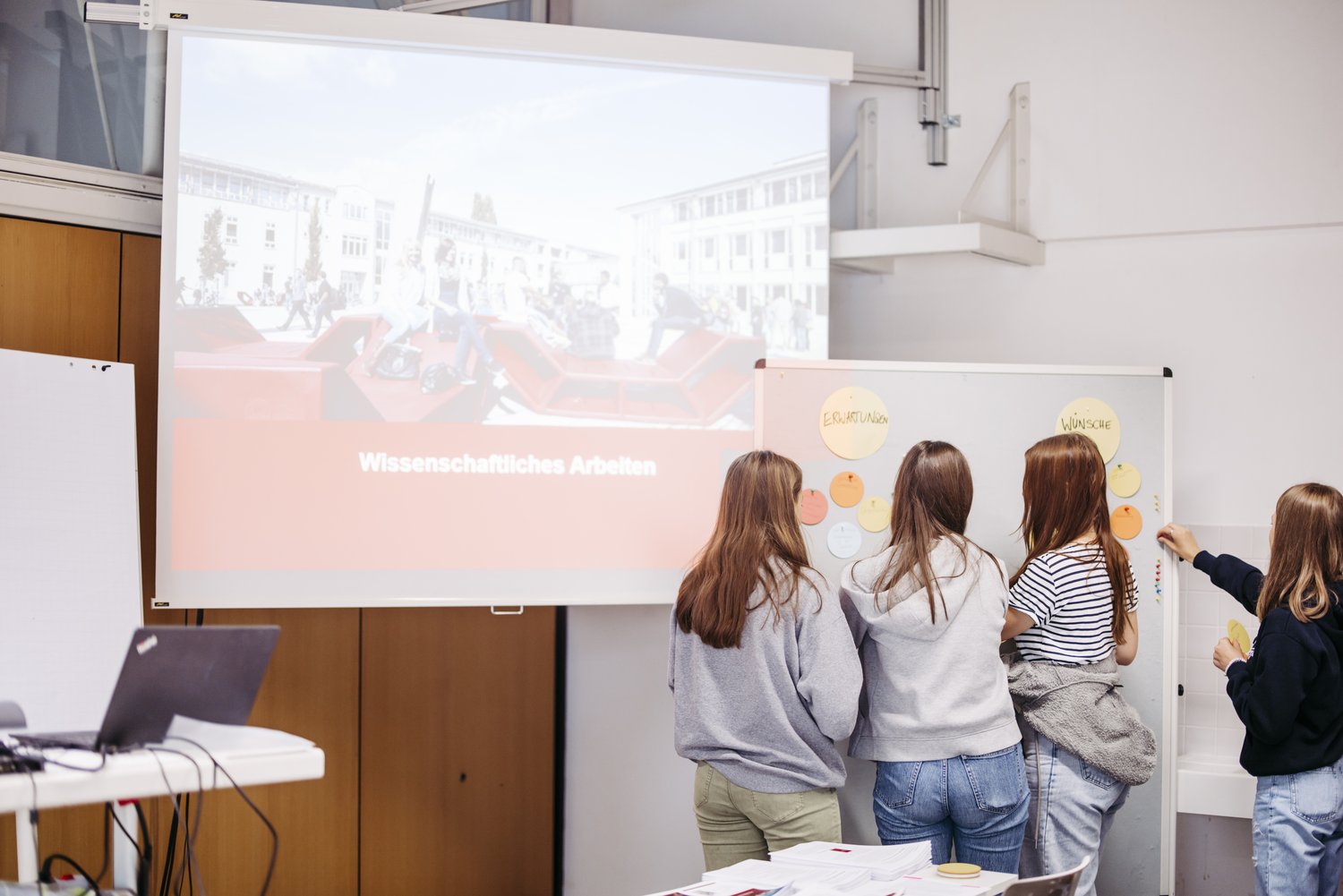 Man sieht drei Personen die vor einem Visionboard. Im Hintergrund sieht man eine große Leinwand.