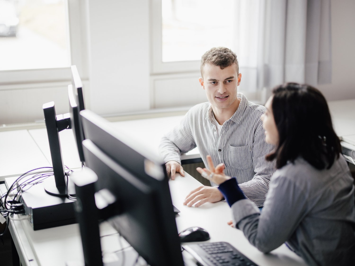 Zwei Personen sitzen an einem Tisch mit Computern und führen ein Gespräch. Die Umgebung ist hell und modern, mit großen Fenstern, die Tageslicht hereinlassen. Beide wirken engagiert und interessiert.