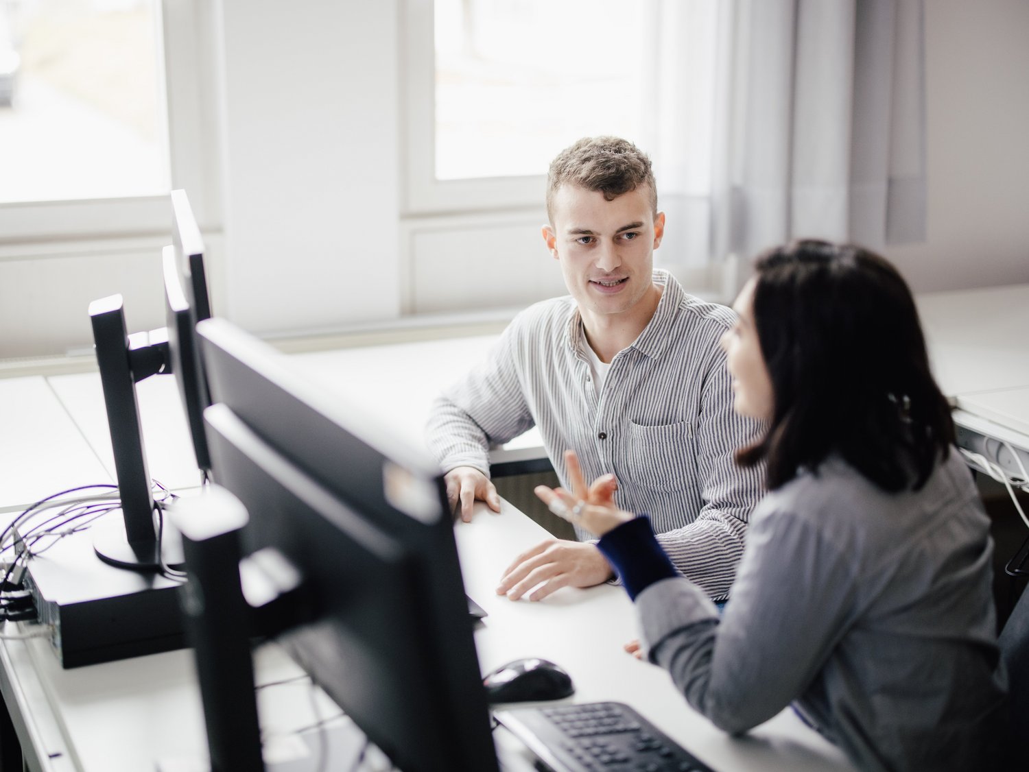 Zwei Personen sitzen an einem Tisch mit Computern und führen ein Gespräch. Die Umgebung ist hell und modern, mit großen Fenstern, die Tageslicht hereinlassen. Beide wirken engagiert und interessiert.
