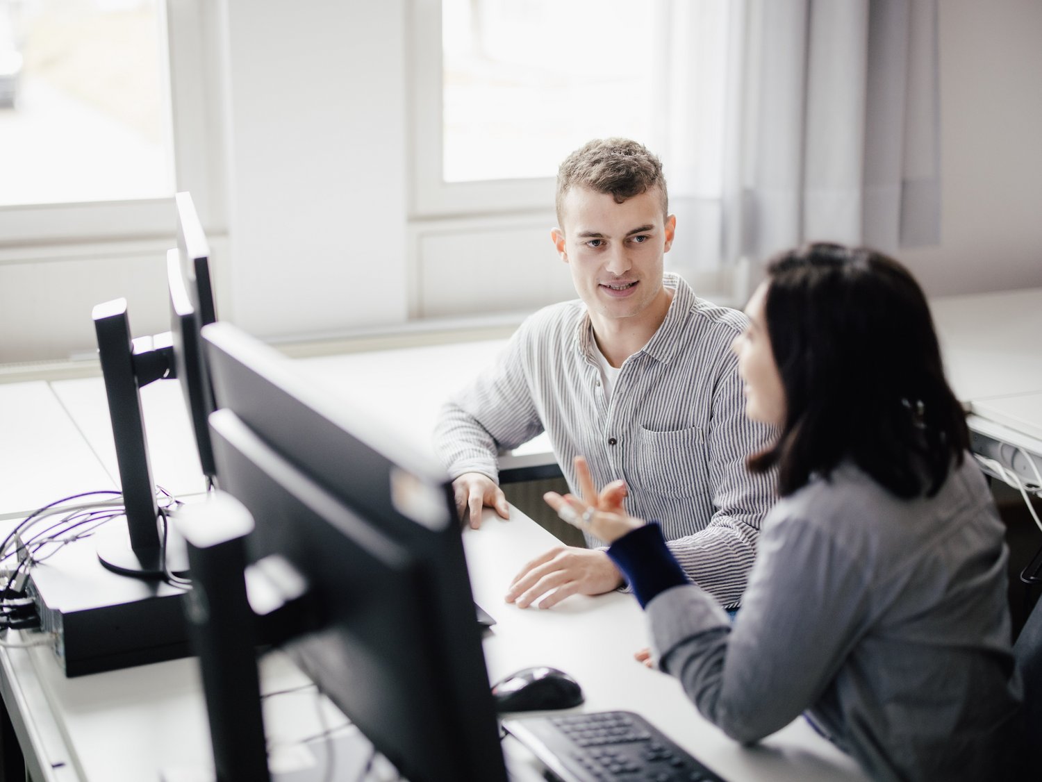 Zwei Personen sitzen an einem Tisch mit Computern und führen ein Gespräch. Die Umgebung ist hell und modern, mit großen Fenstern, die Tageslicht hereinlassen. Beide wirken engagiert und interessiert.