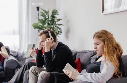 Eine Gruppe von vier Personen sitzt in einem modernen Wohnzimmer. Eine Person trägt Kopfhörer und hört Musik, während die anderen Bücher lesen. Die Atmosphäre ist entspannt und gemütlich, mit Pflanzen und natürlichem Licht im Hintergrund.