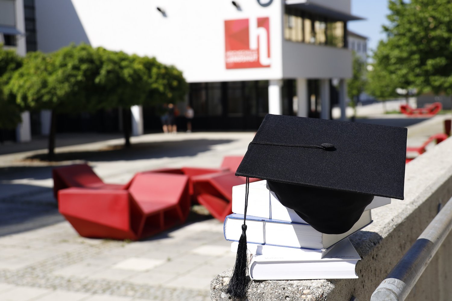 Ein Doktorhut liegt auf Büchern. Im Hintergrund ist die Hochschule Landshut zu sehen.