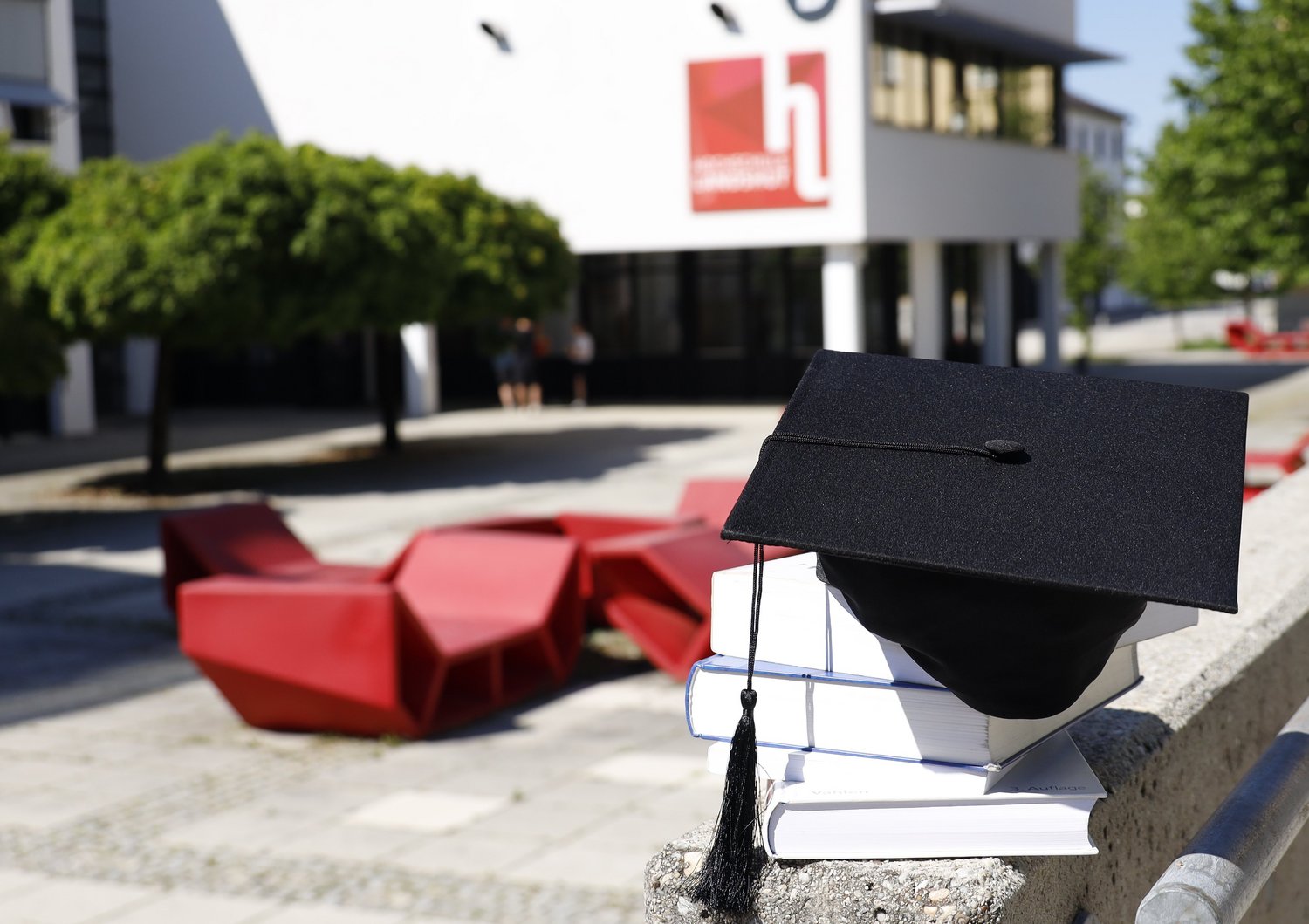 Ein Doktorhut liegt auf Büchern. Im Hintergrund ist die Hochschule Landshut zu sehen.