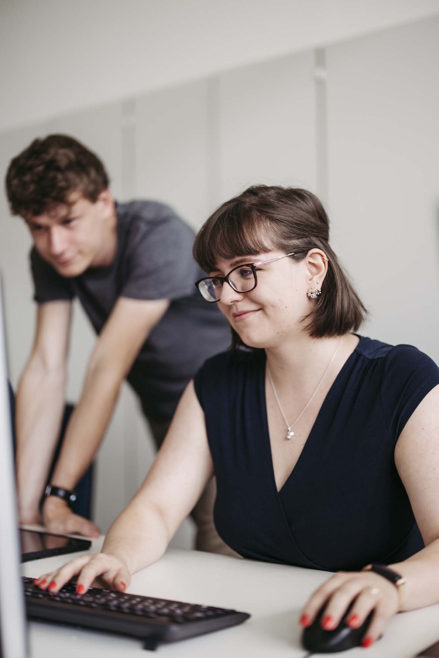 Zwei Personen sind in einem Raum. Die eine Person sitzt und die andere Person steht neben ihr beide schauen in einen Computer.