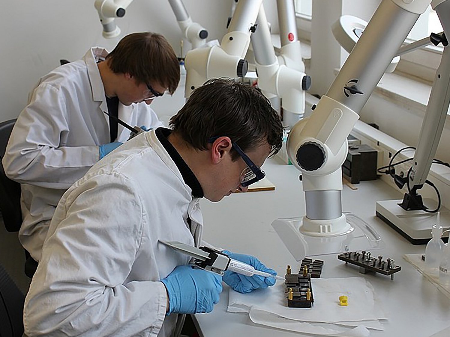 Zwei Personen in weißer Laborkleidung und Schutzbrillen bei der Arbeit mit Spezialklebepistolen am Labortisch