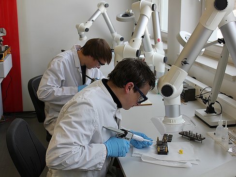 Zwei Personen in weißer Laborkleidung und Schutzbrillen bei der Arbeit mit Spezialklebepistolen am Labortisch
