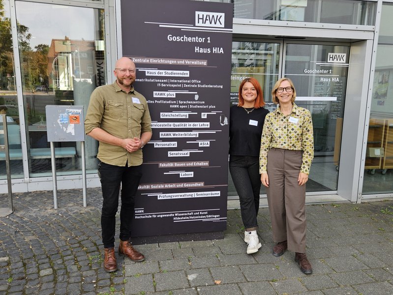 3 Personen stehten vor einem Schild der HAWK