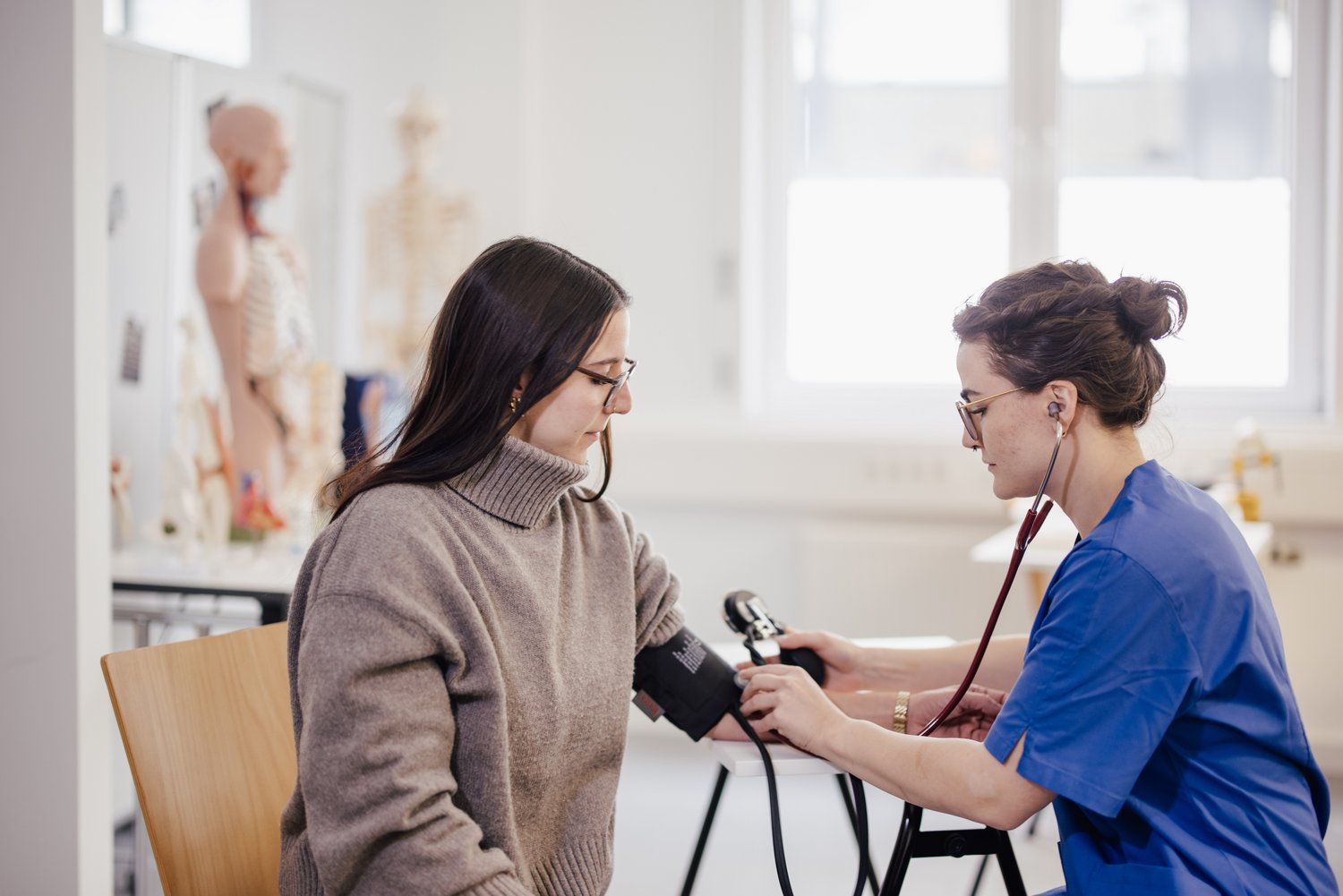 Eine Person sitzt in einem Behandlungsraum und lässt den Blutdruck von einer anderen Person messen. Im Hintergrund sind anatomische Modelle und medizinische Geräte sichtbar.