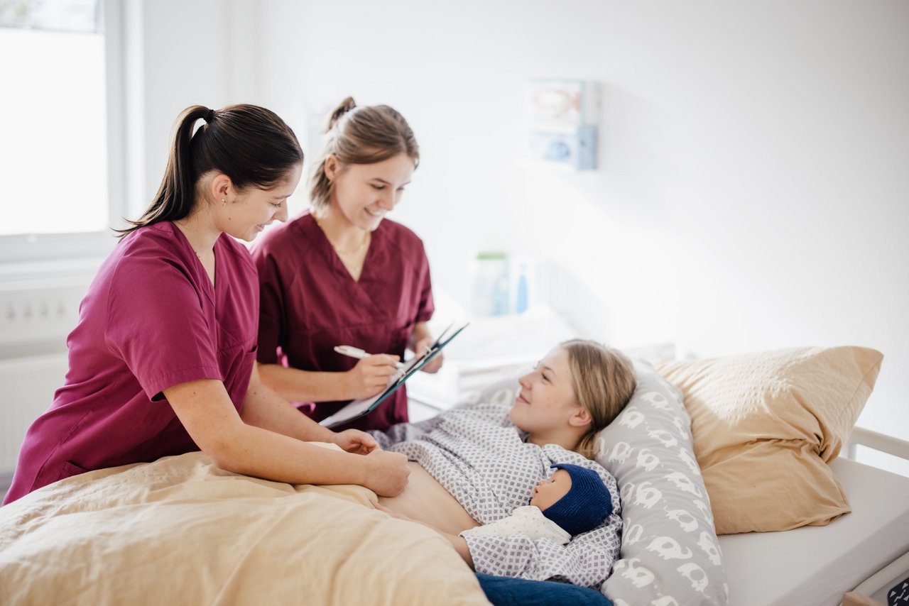 Zwei Pflegekräfte stehen neben einem Bett, während eine Person im Bett liegt. Eine Pflegekraft führt eine Untersuchung durch, während die andere Notizen macht. Der Raum ist hell und freundlich gestaltet.